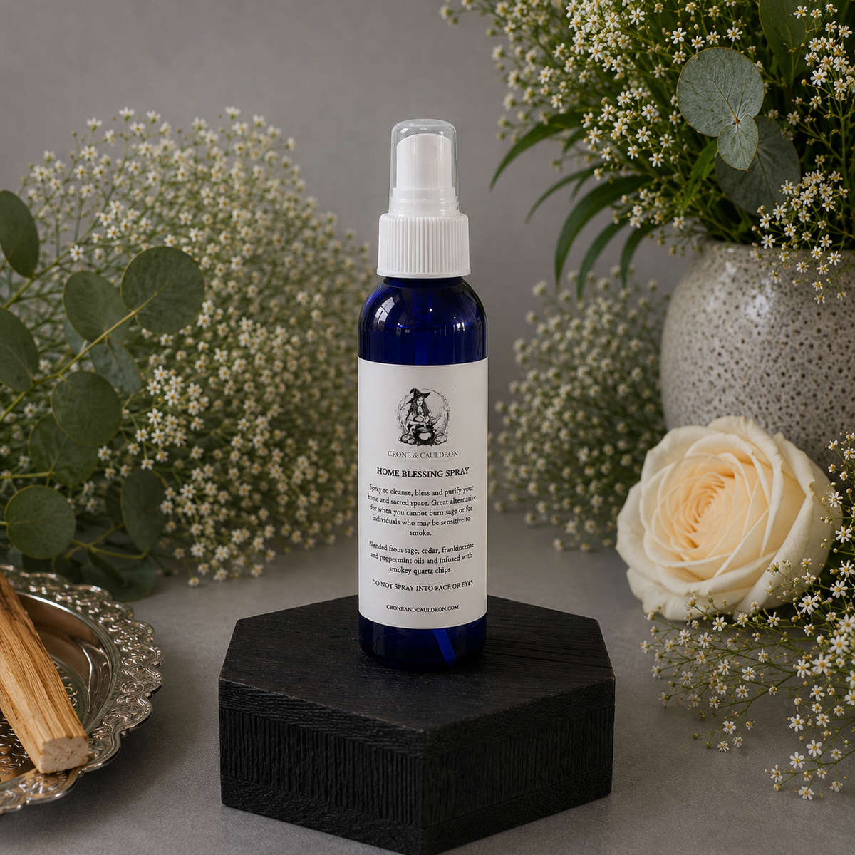 Blue spray bottle with white label on a black stand, surrounded by flowers and plants.