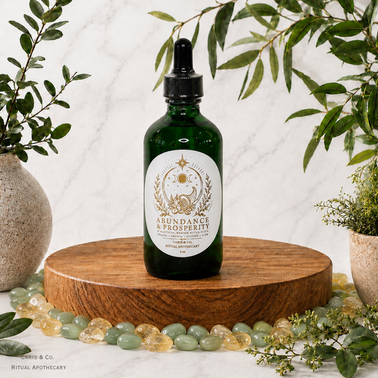 A dark green glass dropper bottle labeled 'Abundance & Prosperity' by  Caris & Co. Ritual Apothecary, displayed on a round wooden pedestal with green leaves, crystals, and ceramic vases around it.