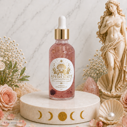 A glass dropper bottle of Aphrodite body oil with pink liquid and gold detailing, displayed on a round marble pedestal with moon phases. The scene is styled with pink roses, a classical statue, and crystals against a marble background.  Caris & Co. Ritual Apothecary