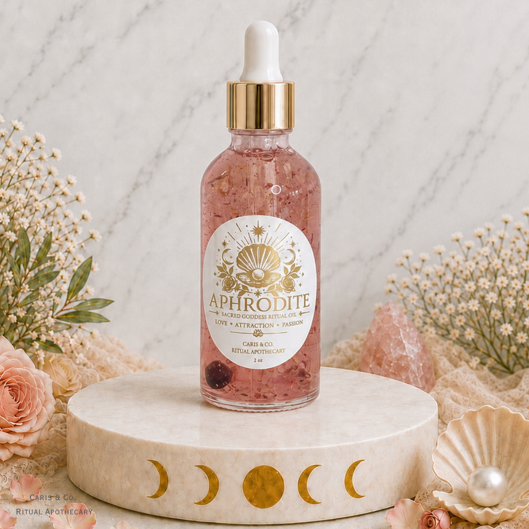 A bottle of Aphrodite Ritual Apothecary rose gold ritual oil with a gold dropper cap, displayed on a round marble pedestal with gold moon phases. The bottle is surrounded by dried flowers, a pink crystal, and a seashell with a pearl against a marble background.  Caris & Co. Ritual Apothecary