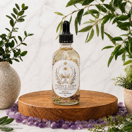 A clear glass dropper bottle labeled 'Crone's Blend' with dried botanicals inside, displayed on a round wooden tray surrounded by purple stones, with green foliage and potted plants against a white marble background.  Caris & Co. Ritual Apothecary