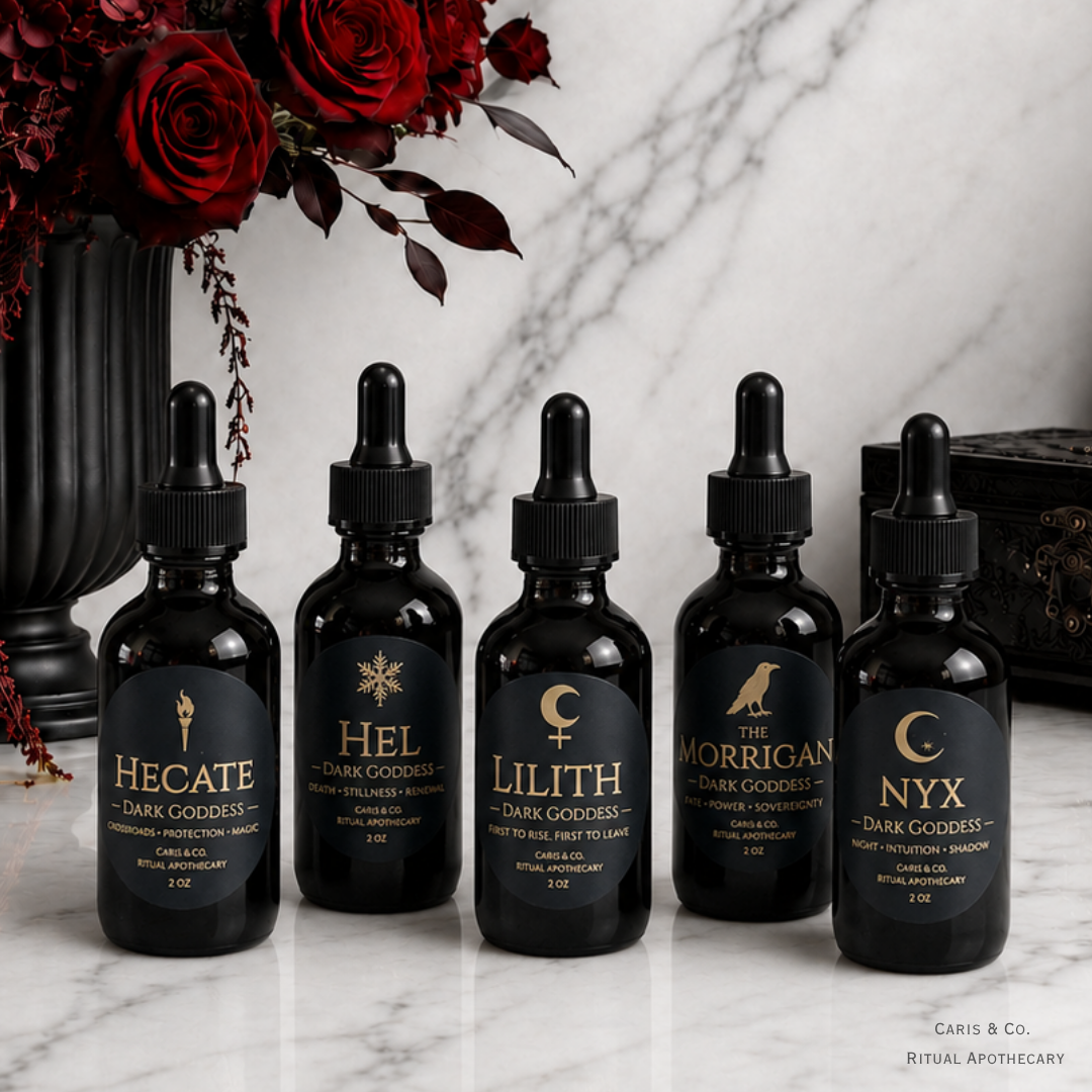 Five black glass dropper bottles labeled Hecate, Hel, Lilith, Morrigan, and Nyx arranged on a white marble surface with red roses in a black vase at the left and a dark wooden box at the right.  Caris & Co. Ritual Apothecary