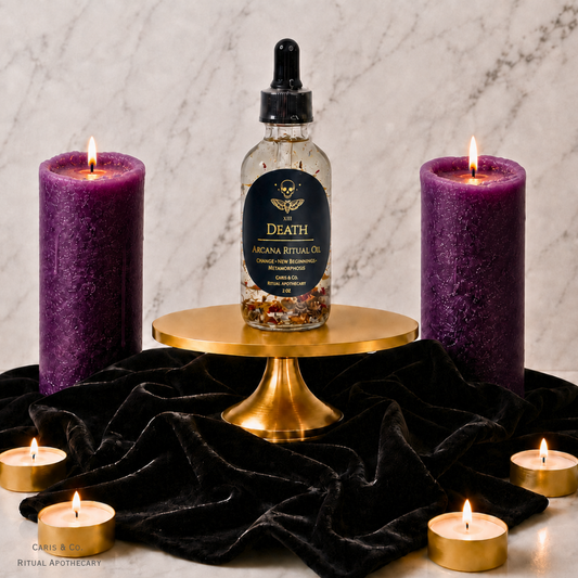 A bottle of Death Arcana Ritual Oil is displayed on a gold pedestal, flanked by two large purple pillar candles and several small tealight candles on a black velvet cloth against a marble background.  Caris & Co. Ritual Apothecary