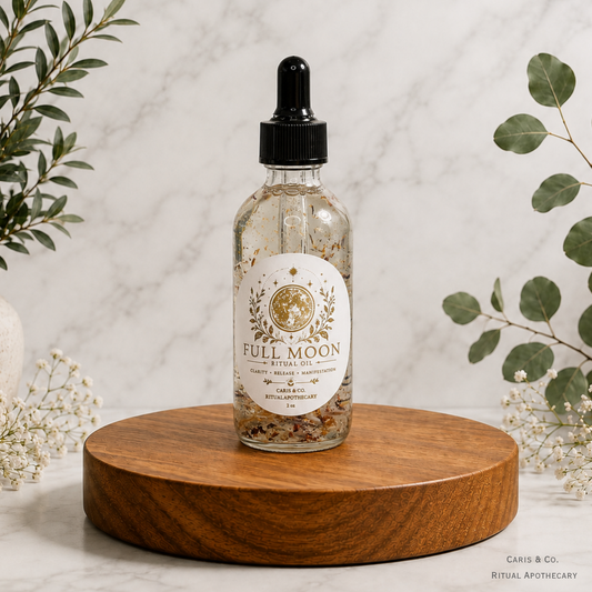A glass dropper bottle of Caris & Co. Full Moon Ritual Oil with dried botanicals, displayed on a round wooden coaster against a marble background with decorative greenery.