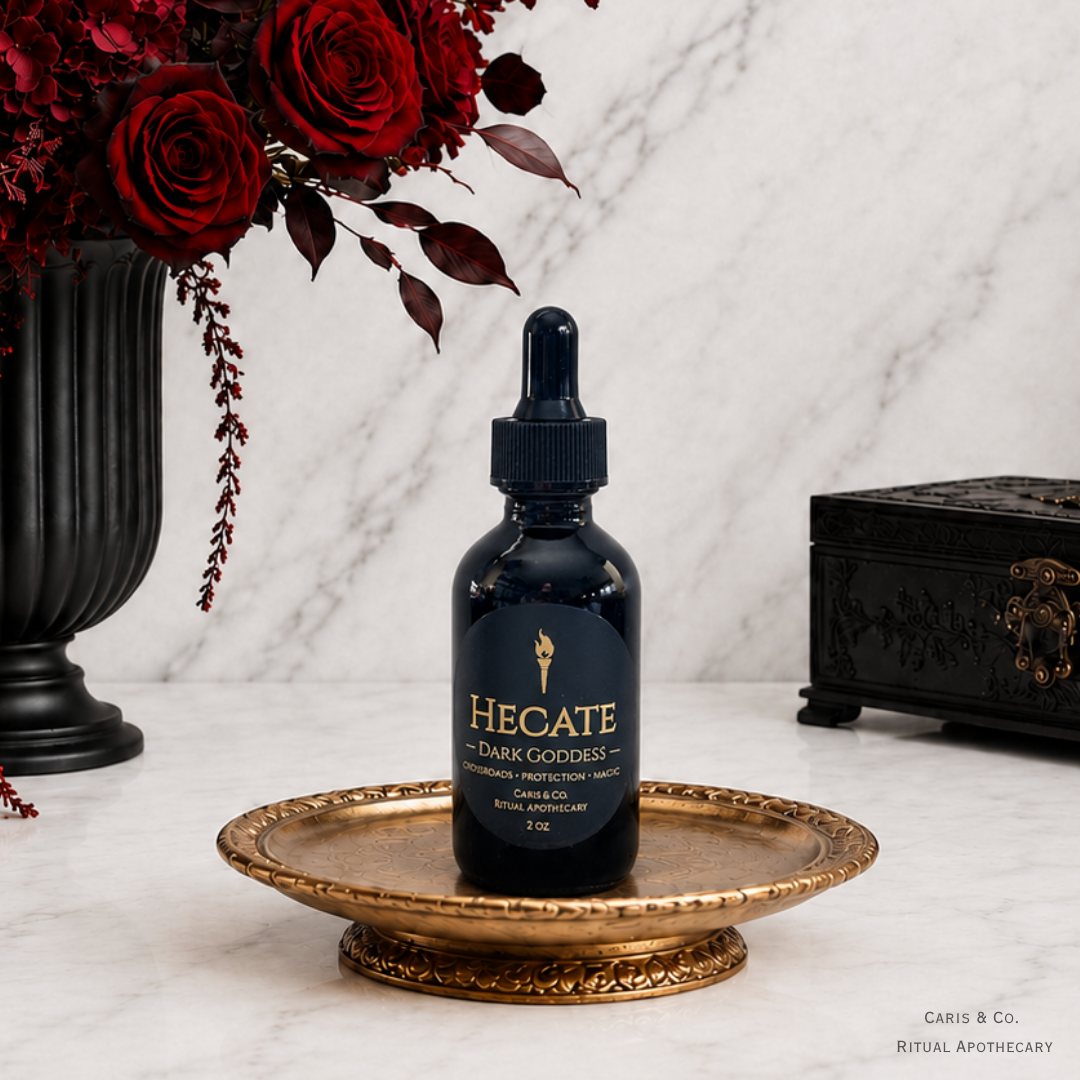 A dark glass dropper bottle labeled 'HECATE Dark Goddess' sits centered on an ornate gold tray on a white marble surface, with red roses in a black vase on the left and a black decorative box on the right. Caris & Co. Ritual Apothecary