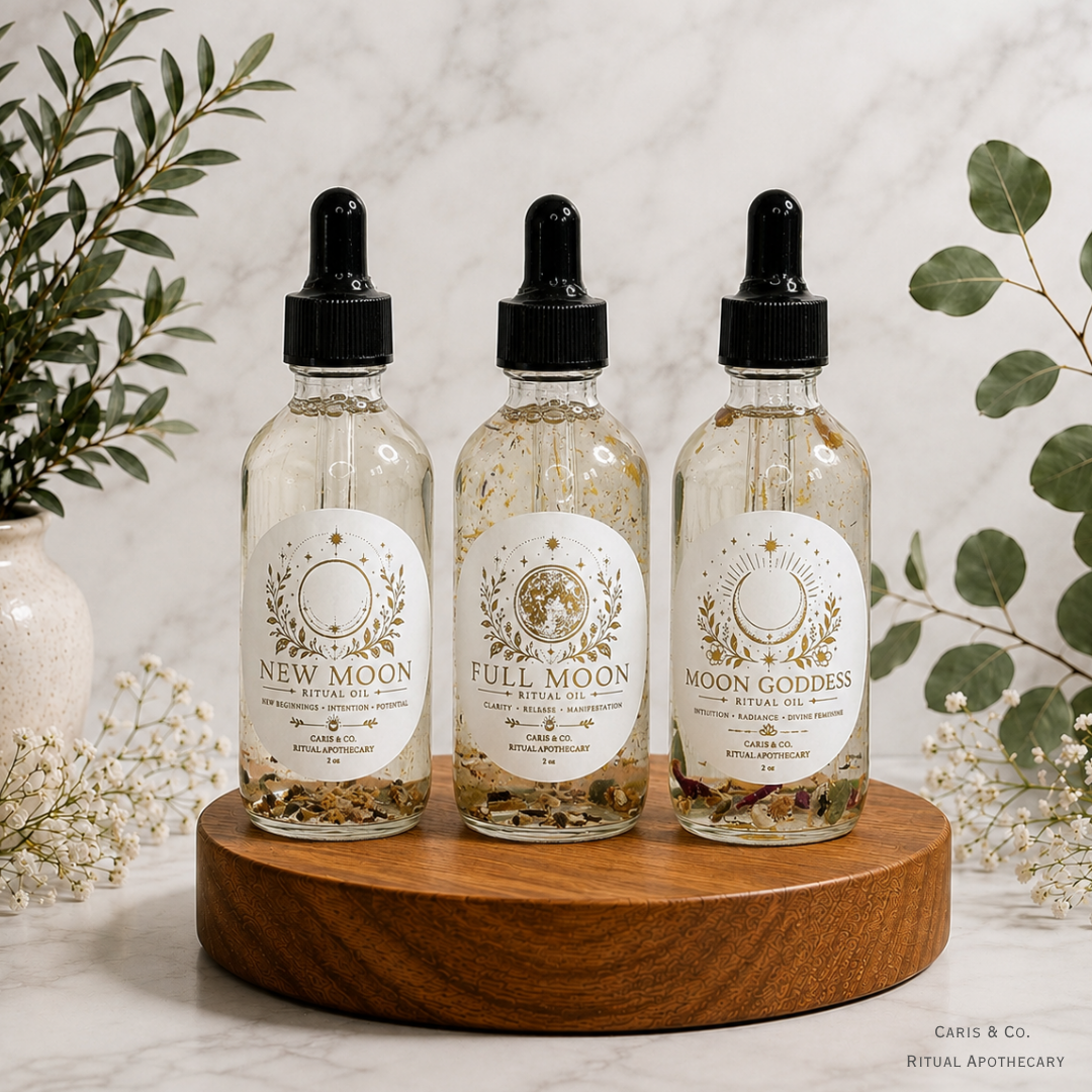 Three glass dropper bottles labeled New Moon, Full Moon, and Moon Goddess, arranged on a round wooden tray with botanical props against a light marble background.  Caris & Co. Ritual Apothecary
