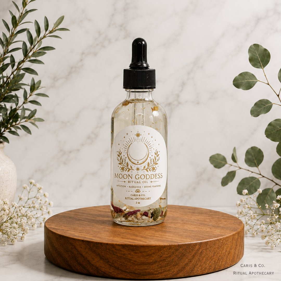 A clear glass dropper bottle of Moon Goddess Ritual Oil with a black cap and white label, displayed on a round wooden pedestal with eucalyptus leaves and baby's breath flowers against a light marble background.  Caris & Co. Ritual Apothecary