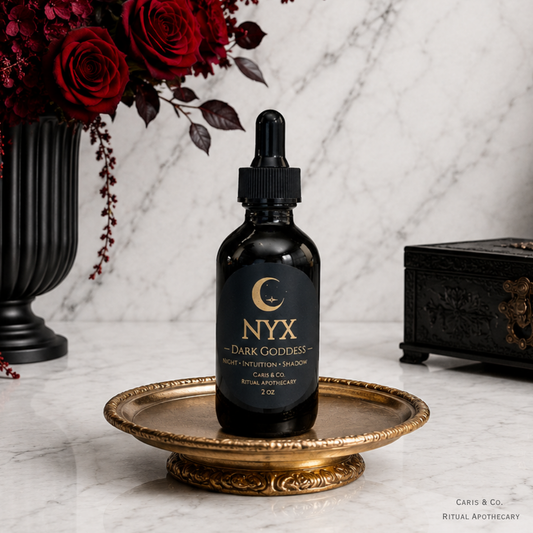 A 2 oz amber glass dropper bottle of NYX ritual oil 'The Dark Goddess' , displayed on an ornate gold tray on a white marble surface with gothic decorative props.  Caris & Co. Ritual Apothecary