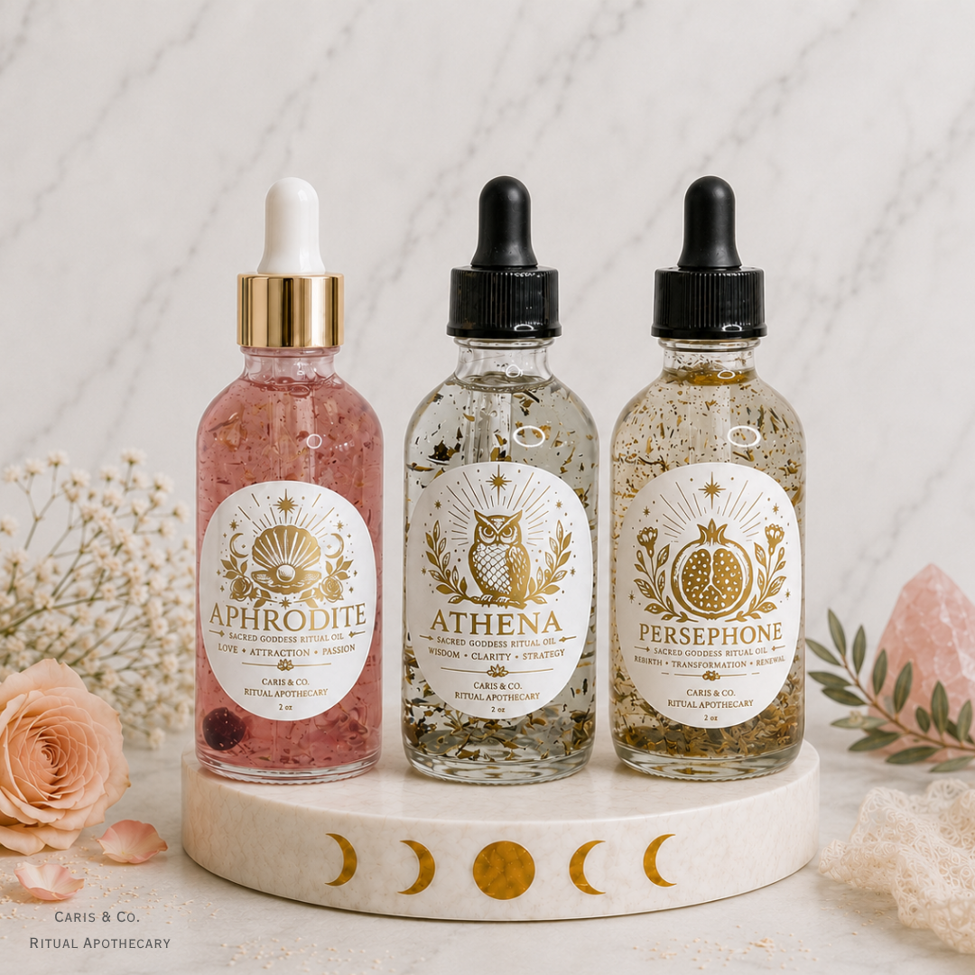 Three glass dropper bottles of Sacred Goddess Ritual Oil by Caris & Co. Ritual Apothecary (Aphrodite, Athena, and Persephone) arranged on a white marble tray with moon phases, surrounded by dried flowers and a rose.