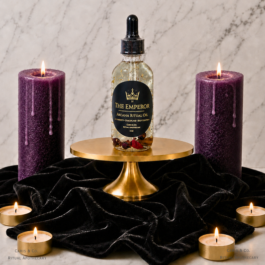 A bottle of The Emperor Arcana Ritual Oil is displayed on a gold pedestal, flanked by two purple wax candles and surrounded by small tealights on a black velvet cloth against a marble background.  Caris & Co. Ritual Apothecary