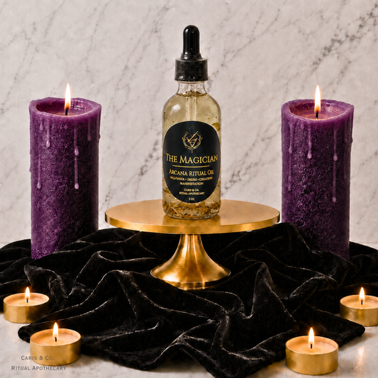 A bottle of The Magician Arcane Ritual Oil by  Caris & Co. Ritual Apothecary sits on a gold pedestal, flanked by two lit purple wax candles and surrounded by small tealight candles on a black velvet cloth against a marble background.