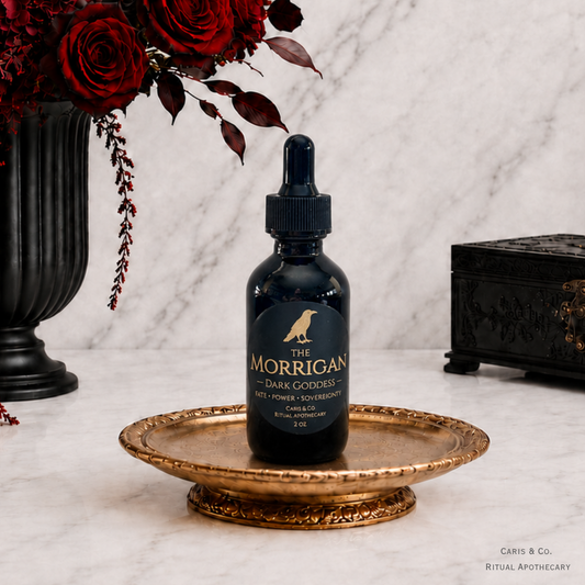A dark glass dropper bottle labeled 'The Morrigan' sits on an ornate gold tray on a white marble surface, styled with red roses in a black vase and a black decorative box in the background. Dark Goddess by  Caris & Co. Ritual Apothecary