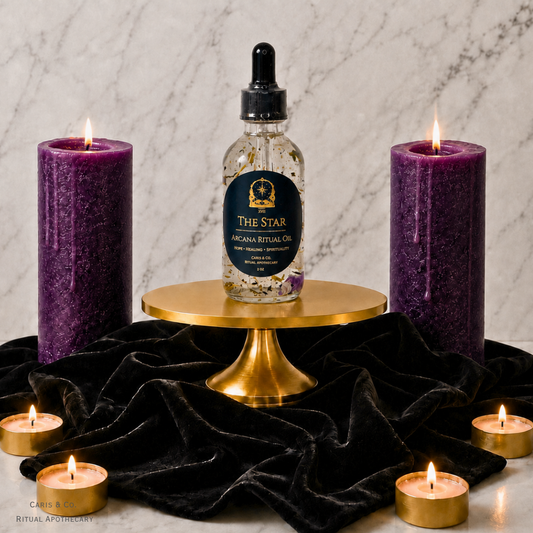 A bottle of The Star Arcana Ritual Oil by  Caris & Co. Ritual Apothecary sits on a gold pedestal, flanked by two lit purple pillar candles and several small tealight candles on a black velvet cloth against a marble background.